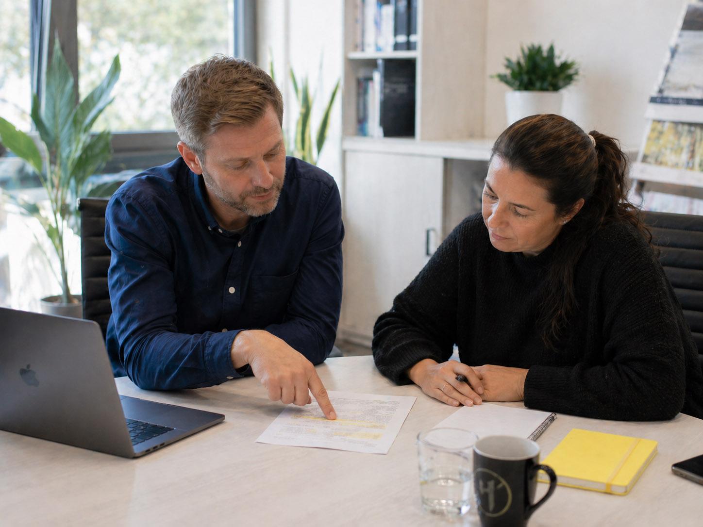 Zwei Personen sitzen an einem Schreibtisch und eine Person zeigt der anderen eine gelbe Markierung auf einem Blatt Papier. Daneben liegt ein gelbes Notizbuch.&nbsp;