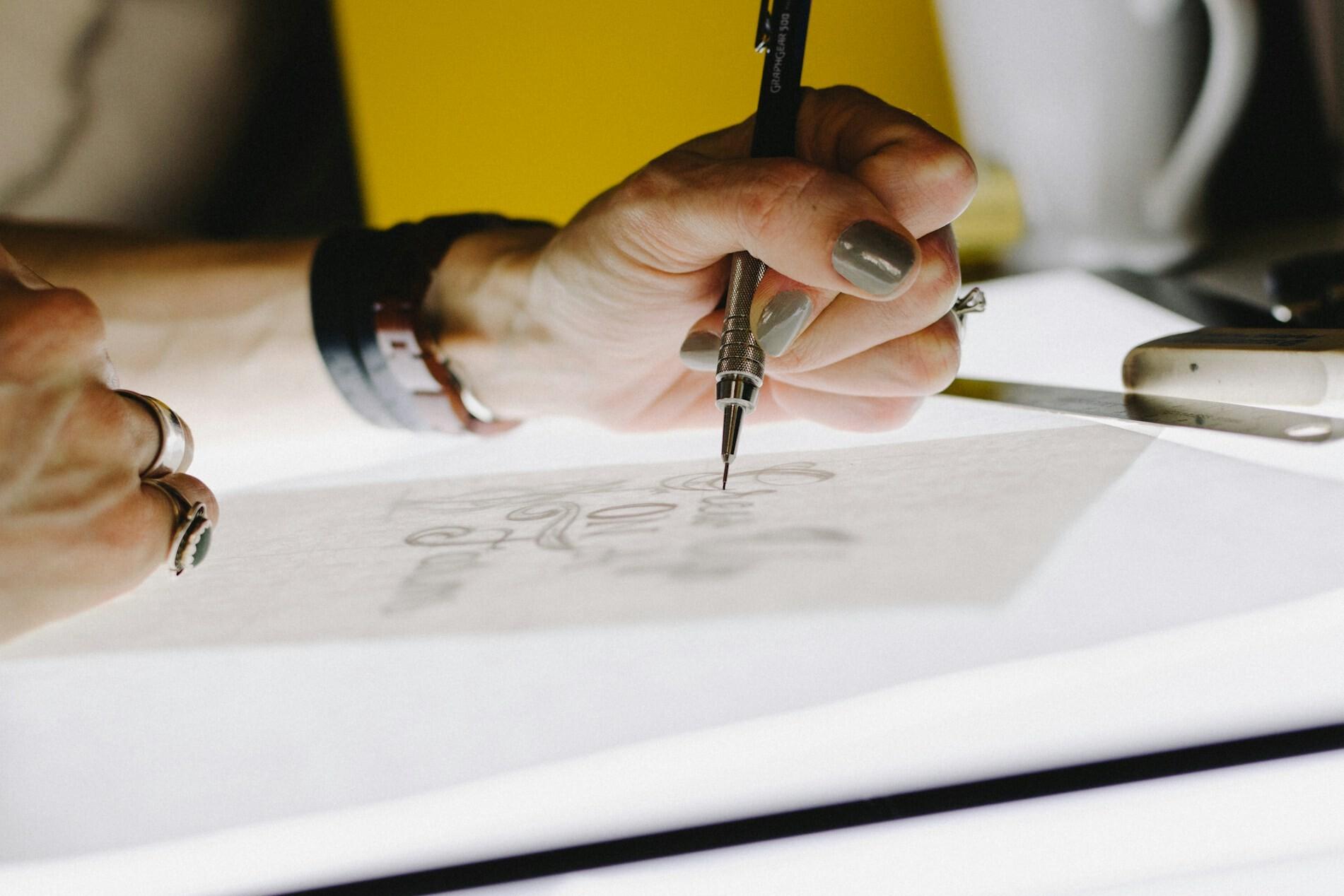 Design Eine Hand mit lackierten Fingernägeln hat einen speziellen Stift in der Hand, mit dem sie Bögen eines Designs nachfährt.