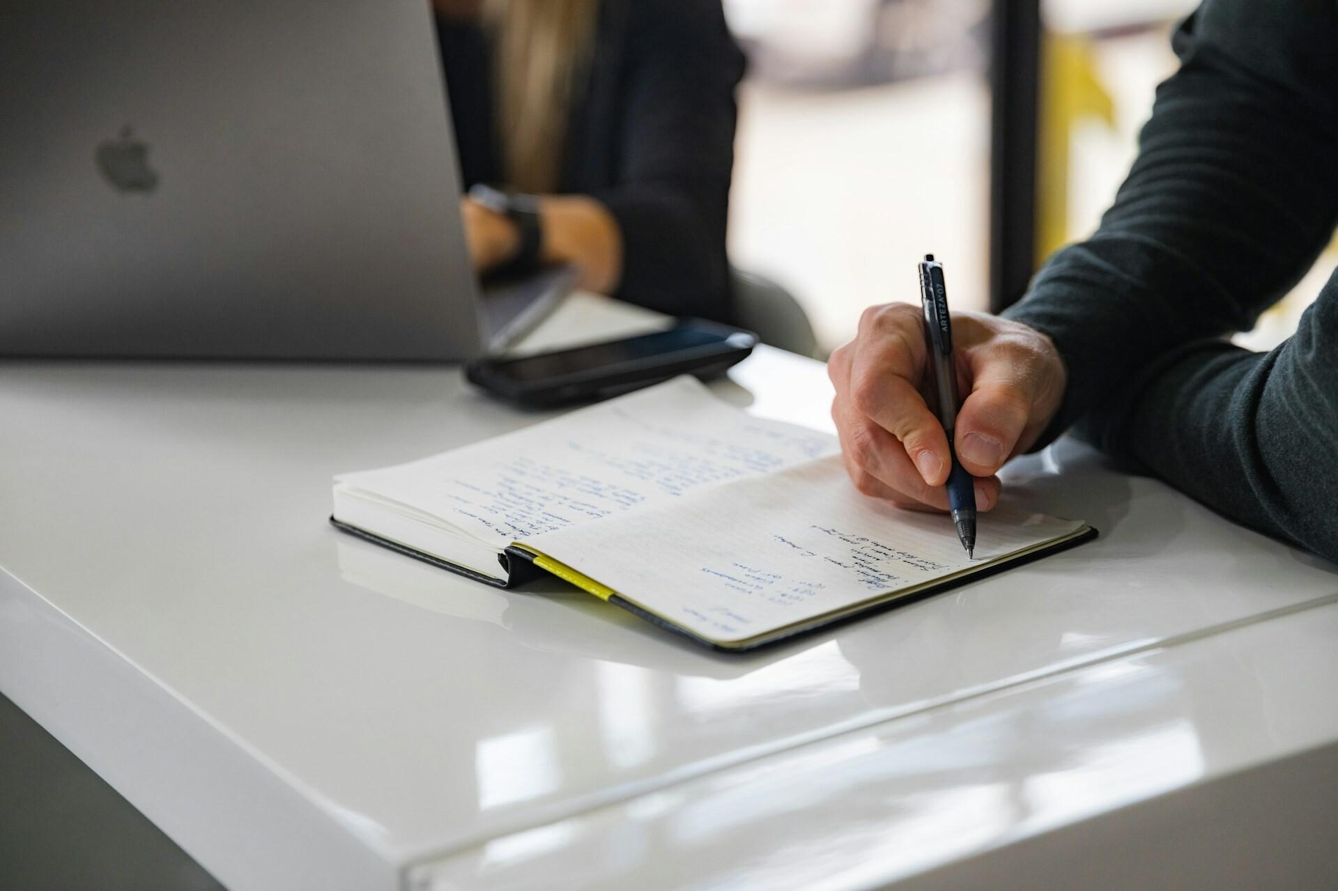 Notizbuch Ein Mann im Anzug, von dem man nur seine Hand sieht, schreibt etwas handschriftlich in ein Notizbuch. Im Hintergrund sieht man eine Frau, die auf einem Laptop tippt. Zwischen ihnen liegt ein Telefon.