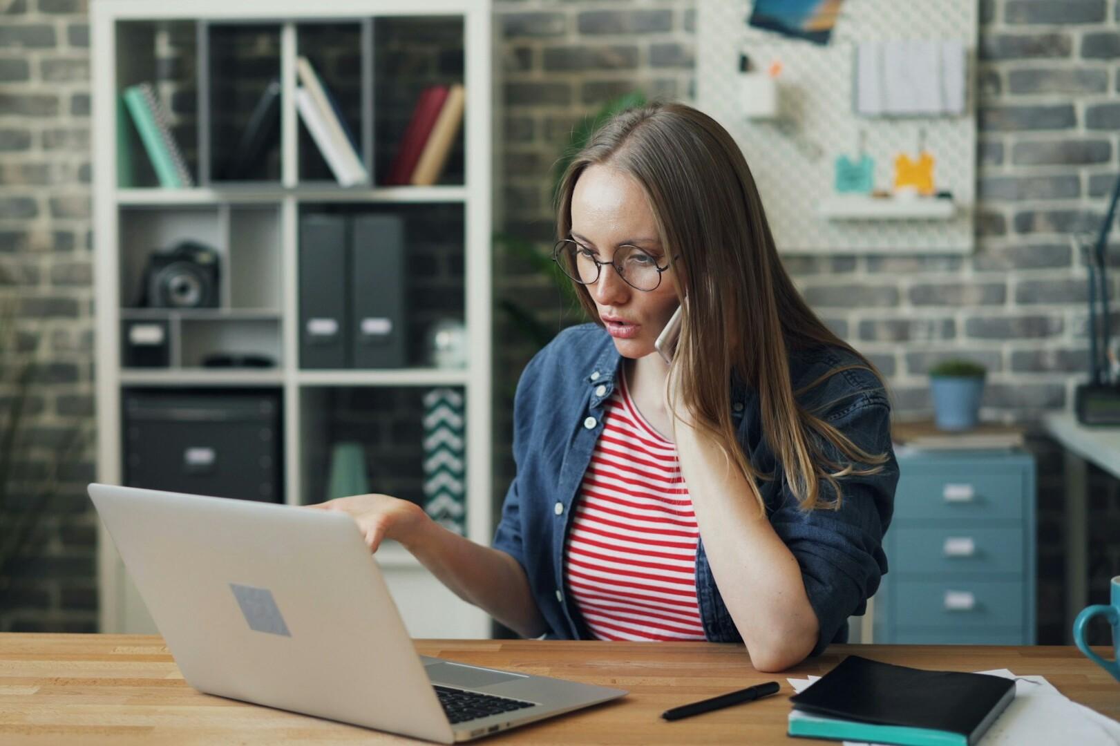 Telefonat Eine Frau telefoniert mit einem Handy an ihrem Schreibtisch und sieht gleichzeitig in ihren Laptop